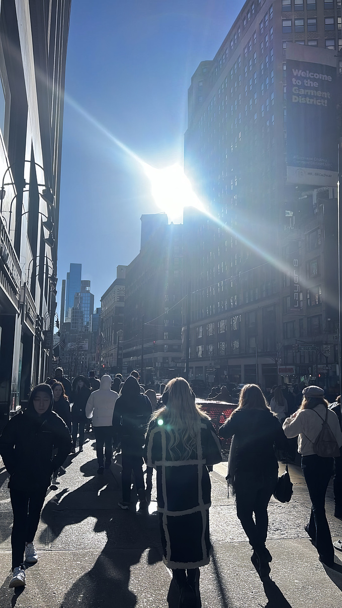 This image captures a bustling street scene in a city of NYC, Manhattan, as I used "Welcome to the Garment District" sign on one of the buildings. The time is early morning, as the sun is low in the sky, casting long shadows across the street.

The street is filled with pedestrians, most of whom are dressed warmly, it's a chilly day. In the foreground, people are walking down the street. The buildings on either side of the street are tall, creating a canyon-like effect, with the sunlight streaming through, adding a dramatic flair to the image. The overall atmosphere feels lively and busy, typical of a city’s daily hustle and bustle.

In this render I tried as much as possible to be realistic, alive and natural.